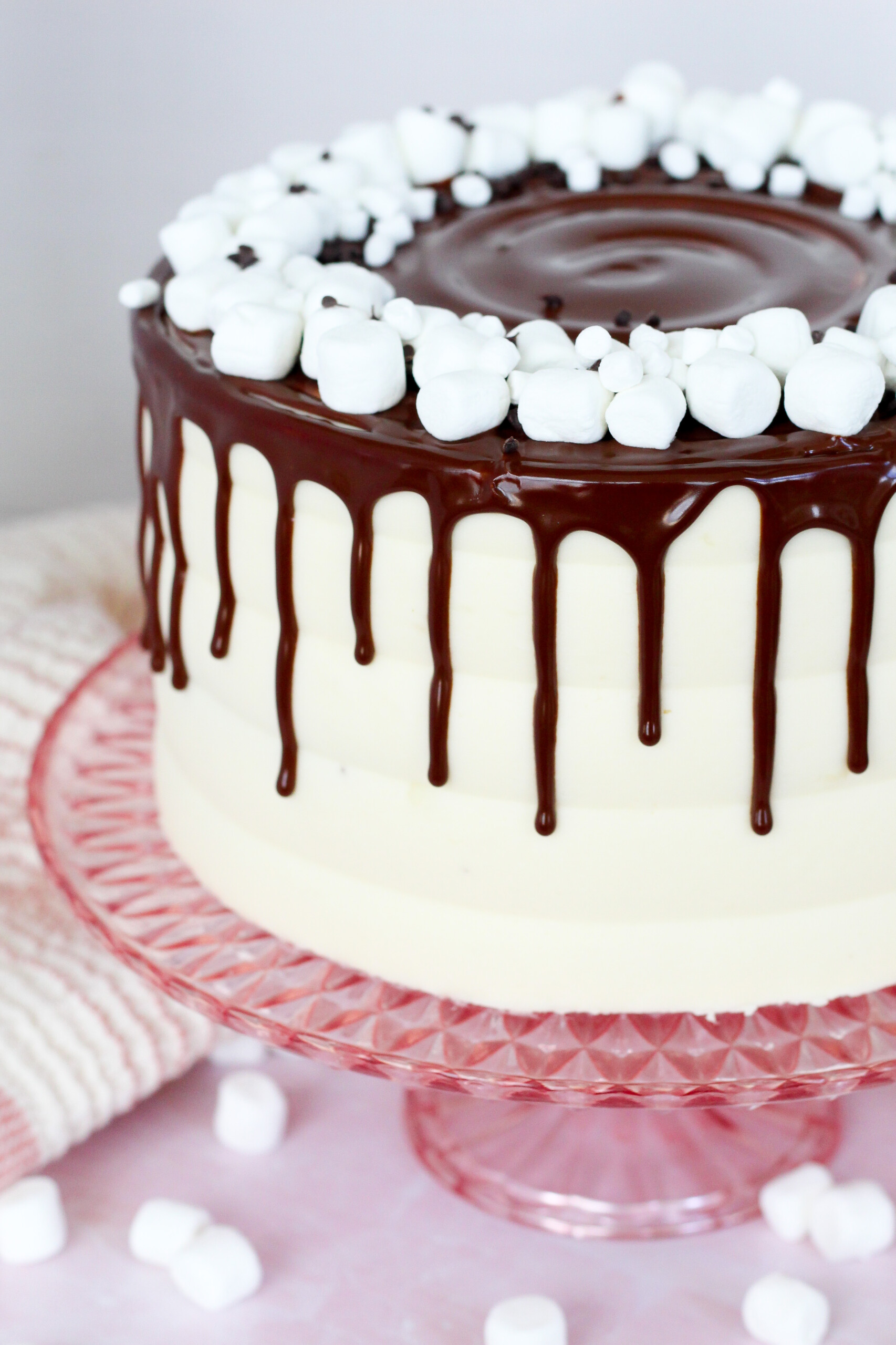 Cake with marshmallow buttercream, a chocolate drip and marshmallows on it, on a cake stand.